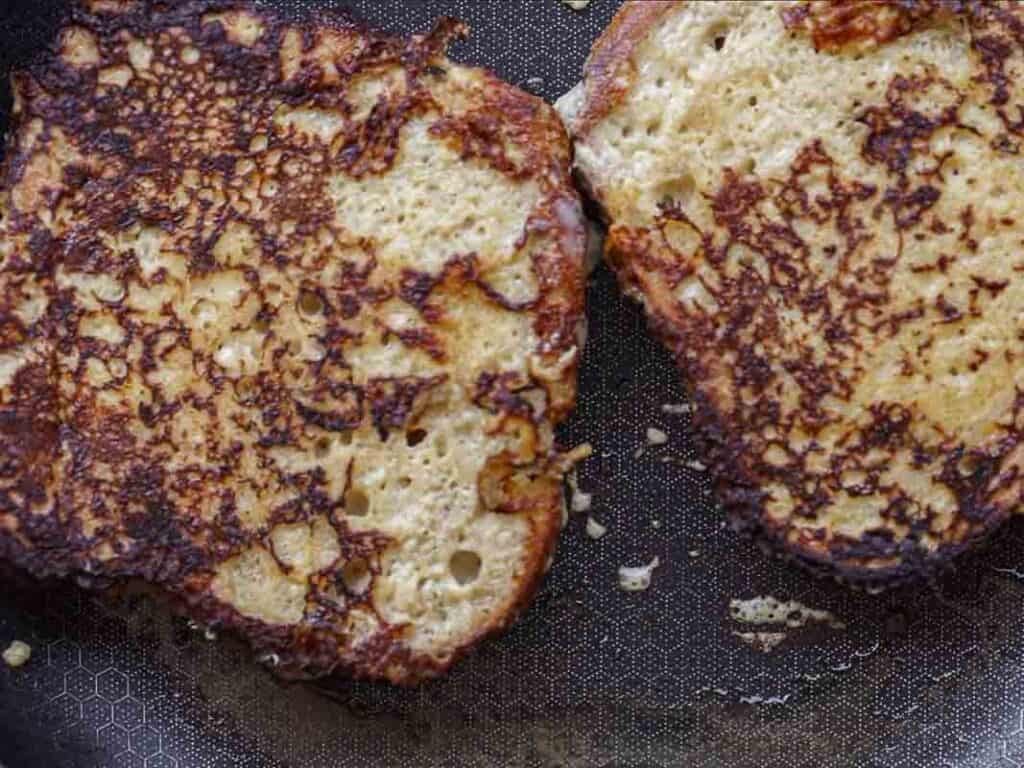 Two slices of vegan French toast are cooking in a skillet, with golden brown, slightly crispy surfaces and some visible bubbles and texture on the bread.